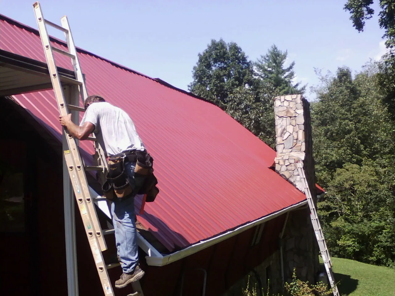 Roofer on a ladder beside a red metal roof and stone chimney — Soffit & Fascia Repair by Roofing Brothers in Minooka